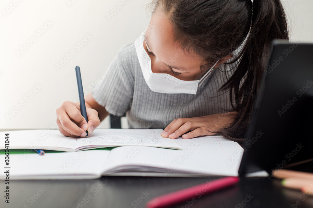 Little girl in a protective face mask does homework for school during quarantine. Distance learning online education during the pandemic of coronavirus Covid-19. Little girl in a protective face mask does homework for school during quarantine. Distance learning online education during the pandemic of coronavirus Covid-19.