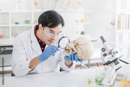 A male scientist with black hair wearing white coat and protective glassware holding a magnifying glass in front of a skull in a white laboratory or hospital.