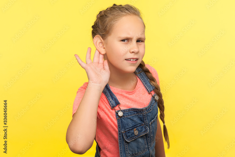 What? I can't hear! Portrait of curious attentive little girl in denim overalls holding hand near ear and listening carefully intently to what you say. indoor studio shot isolated on yellow background What? I can't hear! Portrait of curious attentive little girl in denim overalls holding hand near ear and listening carefully intently to what you say. indoor studio shot isolated on yellow background