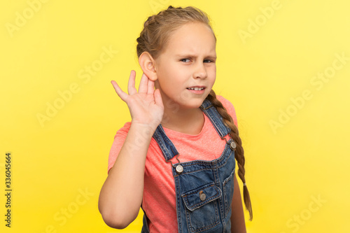 What? I can't hear! Portrait of curious attentive little girl in denim overalls holding hand near ear and listening carefully intently to what you say. indoor studio shot isolated on yellow background
