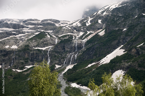 A multi-jet alpine waterfall on a high cliff with a green forest at the foot with glimpses of snow. The concept of a natural landscape in a natural environment on a cloudy day. © yanik88