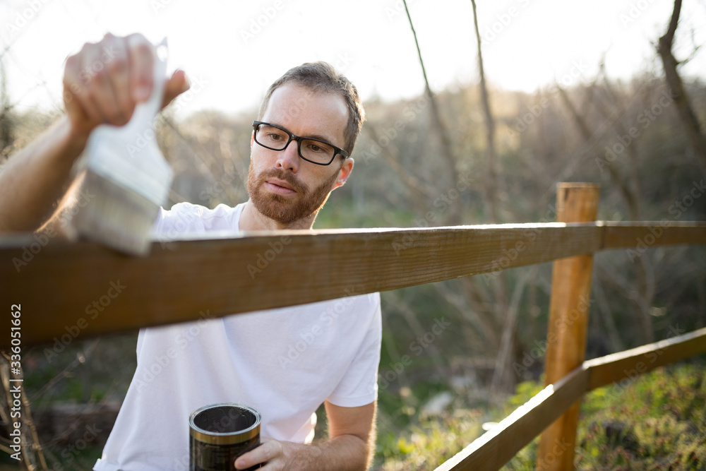 Focused young Caucasian man in casual clothing painting wooden fence in his backyard in late afternoon on a sunny spring day
