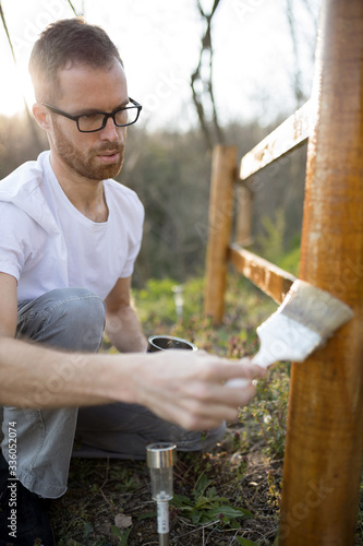 Serious focused young Caucasian man painting wooden fence in his backyard on a beautiful spring afternoon