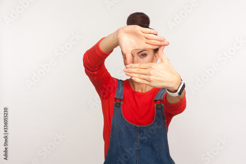 Capture moments. Portrait of girl in denim overalls looking through photo frame shape with fingers, viewing distant, focusing and zooming at camera. indoor studio shot isolated on white background