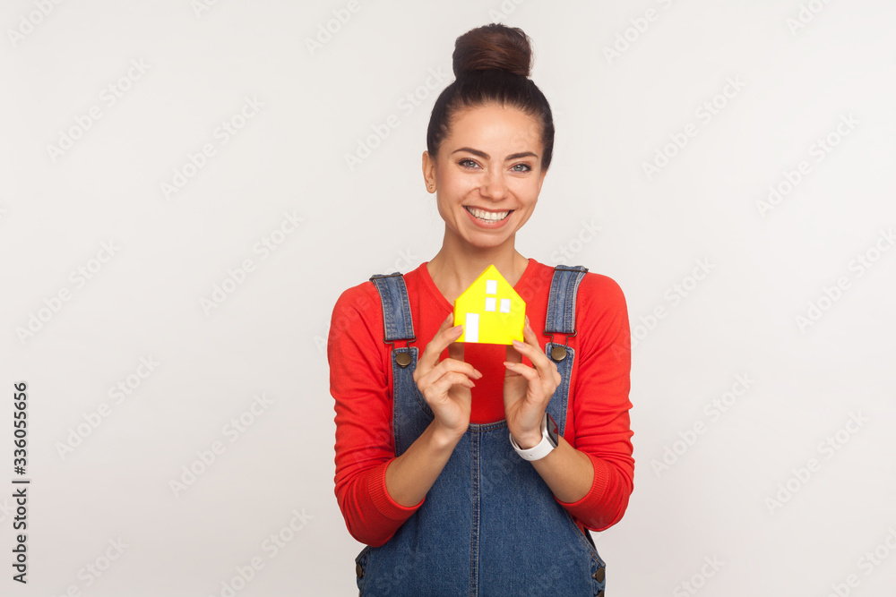 Home purchase. Portrait of charming optimistic girl with hair bun in urban style denim overalls holding paper house and looking at camera with toothy smile. studio shot isolated on white background Home purchase. Portrait of charming optimistic girl with hair bun in urban style denim overalls holding paper house and looking at camera with toothy smile. studio shot isolated on white background