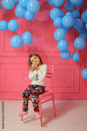 Little girl sitting on chair near many balloons, pink wall in background. photo