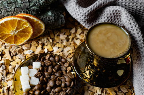 Aromatic dark coffee, with milk with bubbles on the surface in a dark gold Cup on a saucer, next to whole grains, peeled and dried oranges. Background of alder chips and wood snags in the background