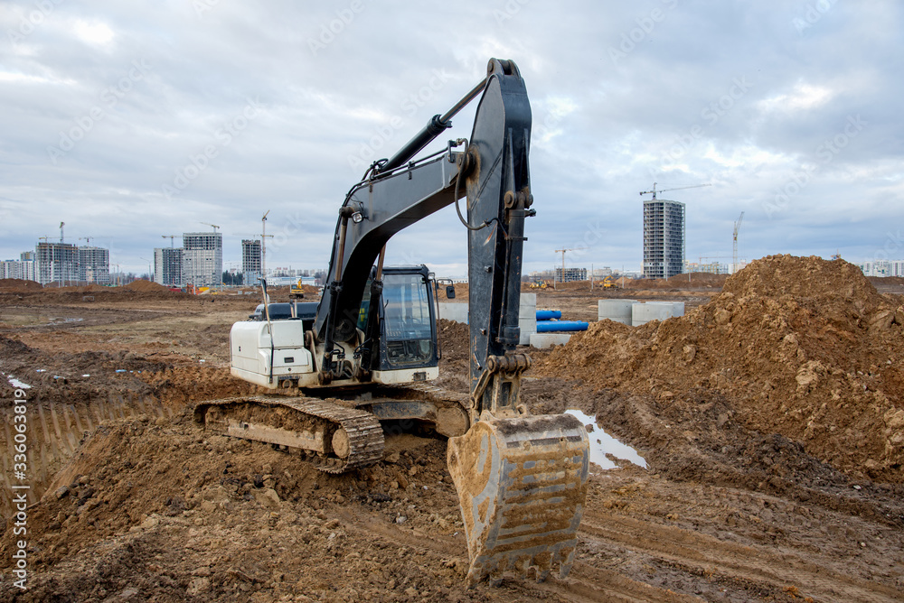 Obraz premium Excavator at earthworks on construction site. Backhoe loader digs a pit for the construction of the foundation. Digging trench for laying sewer pipes drainage in ground. Earth-Moving Heavy Equipment
