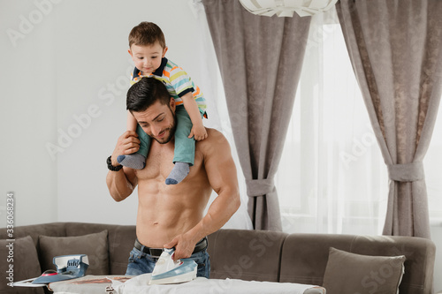 Athletic father ironing a shirt and holding his son on the shoulders. Young fit man at home doing housework.