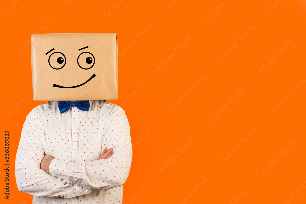 Hombre con caja de cartón en la cabeza con una expresión de alegría en la cara y brazos cruzados sobre fondo naranja liso brillante. Vista de frente. Copy space - obrazy, fototapety, plakaty 