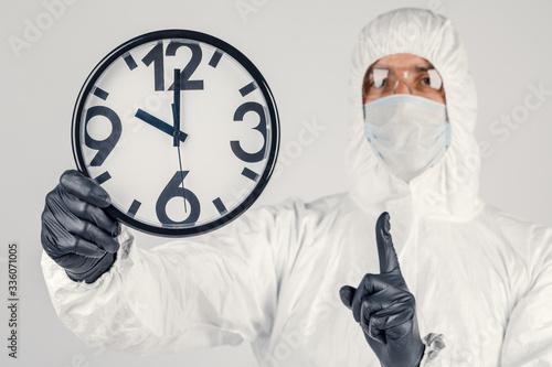 A man in a white protective suit , a mask on a white background, with a watch in his hands. An epidemic pandemic is a new rapidly spreading virus, a medical concept.