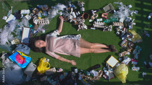 Asian woman among the trash. Top view. An Asian woman lies on the grass among various waste products: glass containers, waste paper, packaging, plastic, disposable, tableware, cans, bottles, boxes.