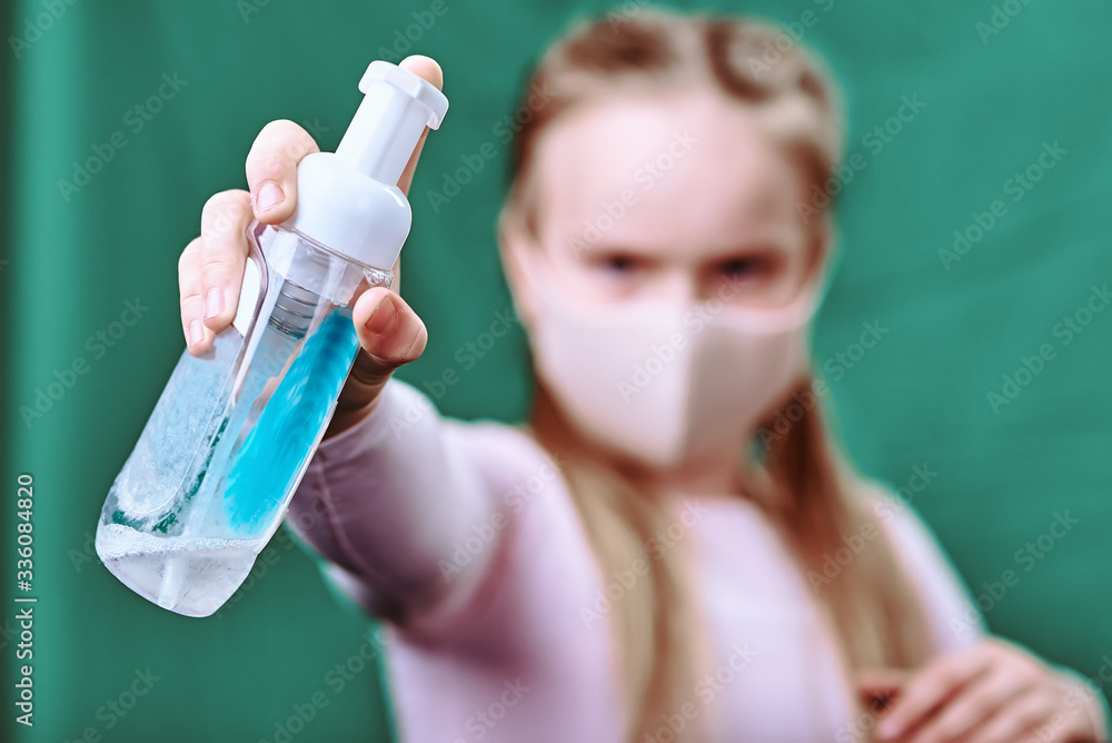 a girl in a medical mask of powder color pours with an antiseptic into a camera. green background. the disinfector in focus. covid-19. to protect health. hygiene and quarantine a girl in a medical mask of powder color pours with an antiseptic into a camera. green background. the disinfector in focus. covid-19. to protect health. hygiene and quarantine