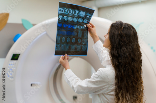 Young caucasian girl doctor looks at x-ray picture of patient brain near kt komputer tomograhphy. Young girl in medical clinic gotes new expirience photo