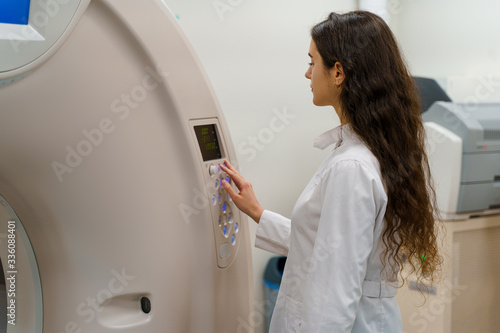 Medical doctor does computer tomography fot patient. Young girl in white coat looks in CT and press button to start procedure research diagnostic