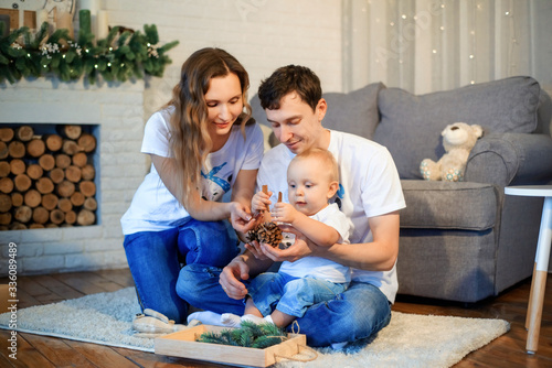 Theme Christmas and New Year family circle. Young Caucasian family with 1 year old toddler son sitting on floor in living room. Christmas decorations. Boy and parents. Family self isolation at home.