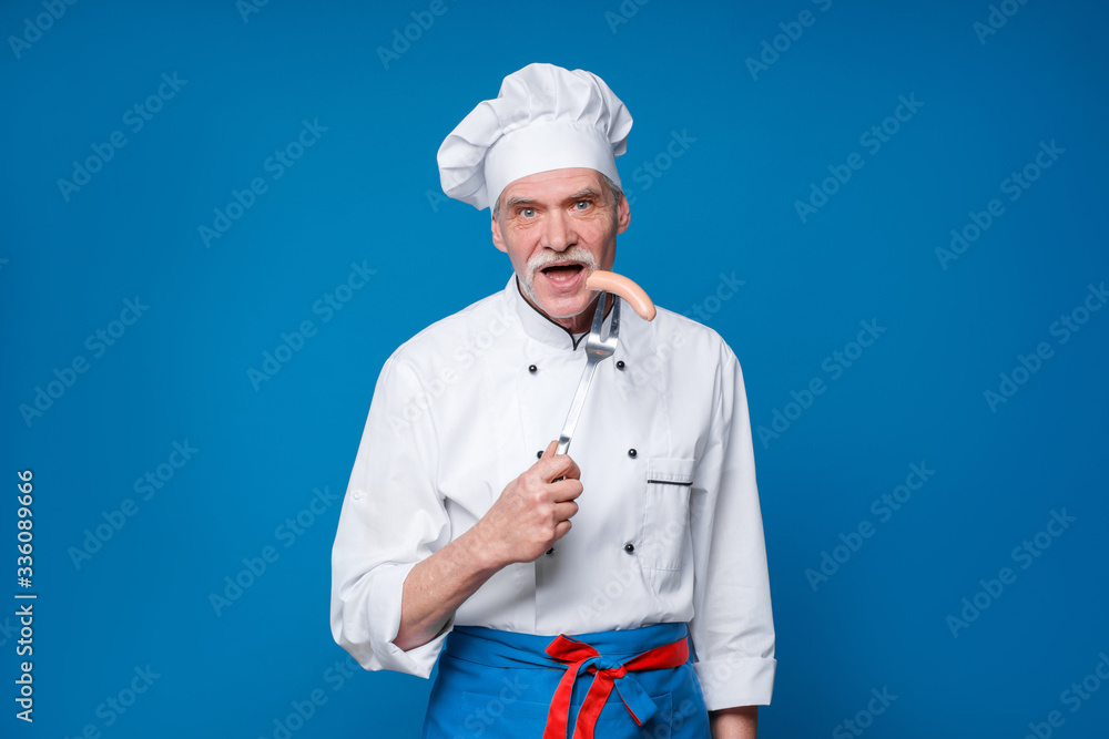 Portrait of a smiling senior chef holding sausage on special fork, isolated on blue background Portrait of a smiling senior chef holding sausage on special fork, isolated on blue background