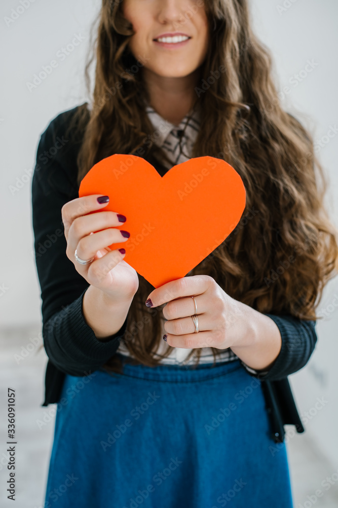 happiness, health, people, holidays and love concept - smiling young woman in white t-shirt holding red heart against the window happiness, health, people, holidays and love concept - smiling young woman in white t-shirt holding red heart against the window