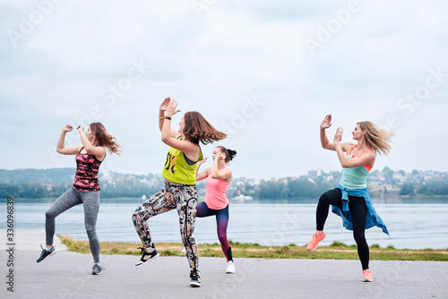 A group of young women, wearing colorful sports outfits, doing zumba exercises outside by city lake. Dancing training to loose weight in summer. Healthy lifestyle concept. Female sport leisure. photo