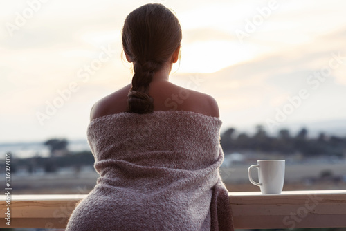 Beautiful alone nude brunette woman wrapped in a plaid on the balcony of her bedroom with a cup of coffee in the early morning © flowertiare