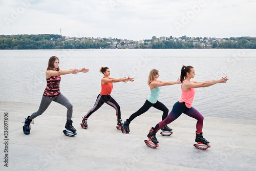 A group of young fit slim women in kangoo jumps, training in front of city lake in summer. Four girls, wearing colorful sports outfit, doing exercises outdoors. Healthy lifestyle concept. © Natalia
