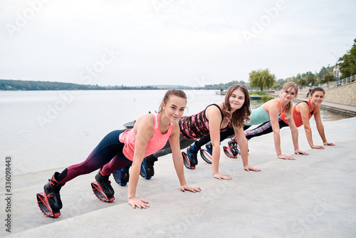 A group of young fit slim women in kangoo jumps, training in front of city lake in summer. Four girls, wearing colorful sports outfit, doing exercises plank outdoors. Healthy lifestyle concept.