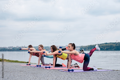 A group of four young women, wearing colorful sports outfit,doing fitness exercises on yoga mats outside by city lake in summer. Workout power female training to loose weight and body shape at nature. © Natalia