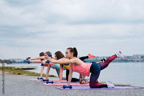 A group of four young women, wearing colorful sports outfit,doing fitness exercises on yoga mats outside by city lake in summer. Workout power female training to loose weight and body shape at nature.