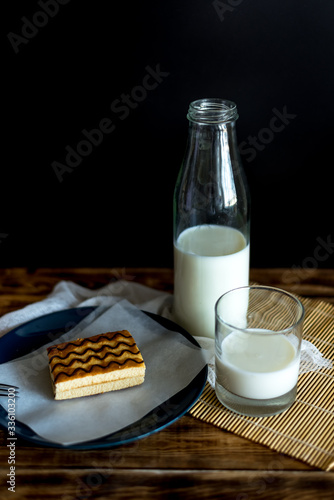 on a black background on a wooden table is a glass bottle with milk, next to a glass of milk and a cake