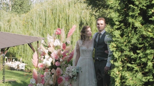 The bride and groom stand against the background of the wedding arch on a summer day, made of fresh flowers and pampan grass, medium-sized and smile photo