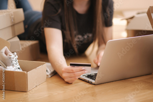 Young woman doing online shopping at home. Female lying on the wooden floor with different parcel. Woman using laptop and credit card © Aleksandr