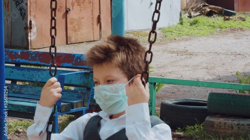 Young handsome lonely student in protective medical mask on his face rides with sad on a swing, hold rusty metal chains, looks alone around by during coronavirus isolation period on sunny day close up photo