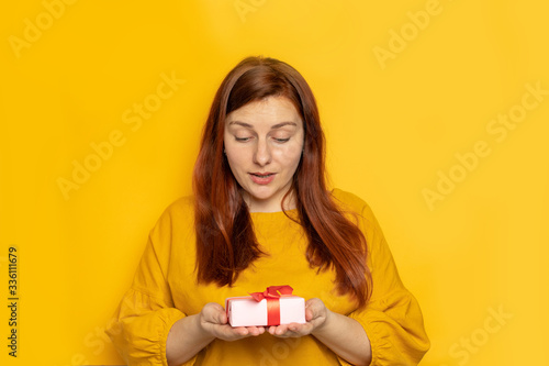 Cheerful woman holds a gift box in her hands and opens a gift on a yellow background wall. Giving Day, Womens Day, Saint Valentine's Day concept. She expresses joy.