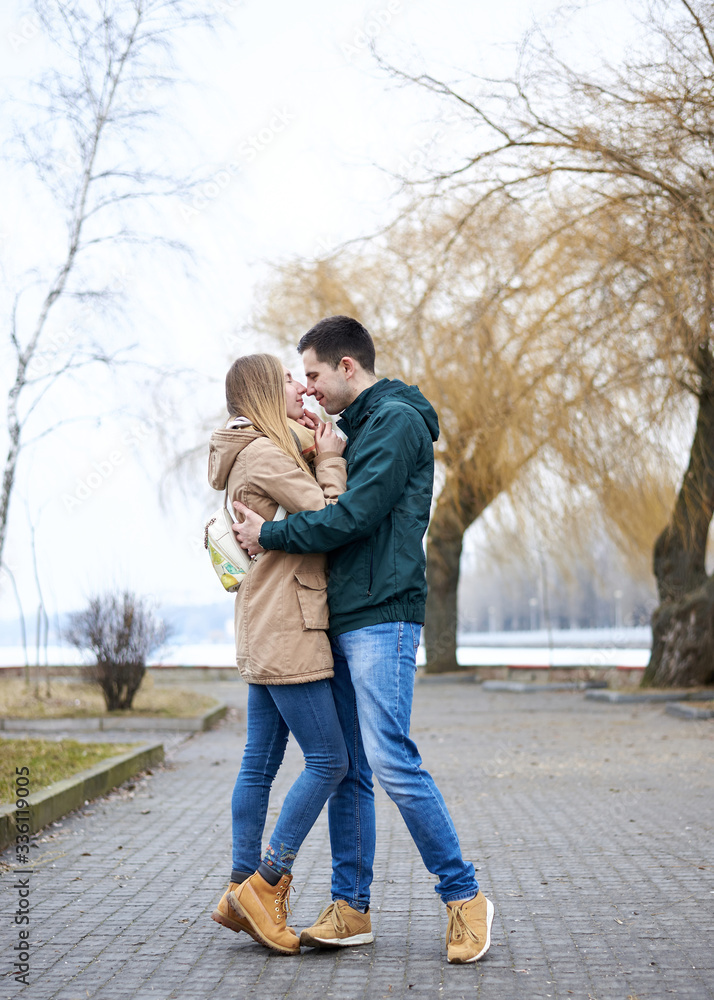 Young couple in love, standing, embracing hugging each other, wearing casual clothes and jeans, on the rainy spring day in city park. Romantic Valentines day celebration outside in town. Young couple in love, standing, embracing hugging each other, wearing casual clothes and jeans, on the rainy spring day in city park. Romantic Valentines day celebration outside in town.