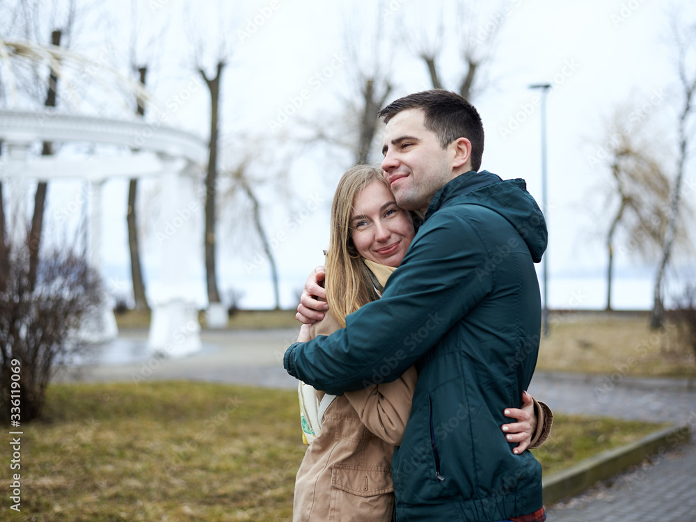Young couple in love, standing, embracing hugging each other, wearing casual clothes and jeans, on the rainy spring day in city park. Romantic Valentines day celebration outside in town. Young couple in love, standing, embracing hugging each other, wearing casual clothes and jeans, on the rainy spring day in city park. Romantic Valentines day celebration outside in town.