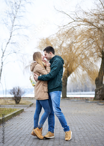 Young couple in love, standing, embracing hugging each other, wearing casual clothes and jeans, on the rainy spring day in city park. Romantic Valentines day celebration outside in town.
