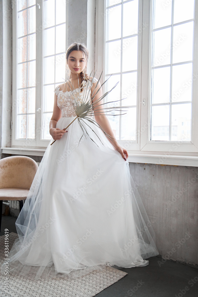 Bride is a woman in a light summer wedding dress standing at the window. Brown-haired girl with perfect hair and beautiful makeup