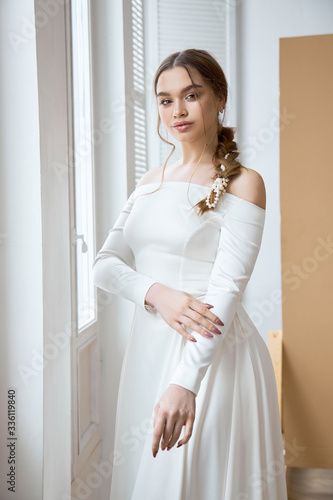 Beautiful slender blonde in the evening sun in a long white dress near a large window. Perfect hair and makeup for the bride, a new collection of wedding dresses