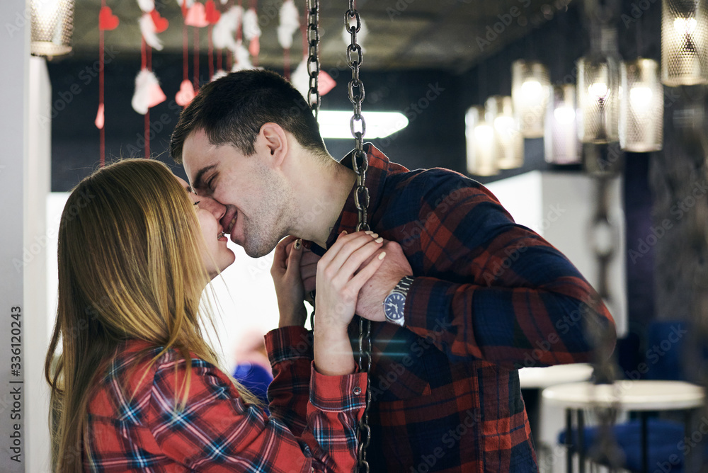 Young couple in love, wearing red checkered shirts, sitting on a swing in coffee shop. Attractive brunette man kissing pretty blond woman in coffee shop. Romantic Valentines day celebration