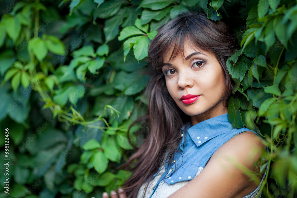 Close up of attractive brunette girl posing among green leaves. Charming lady with raspberry lipstick on lips looking at camera and smiling slightly. Concept of female beauty and nature. Close up of attractive brunette girl posing among green leaves. Charming lady with raspberry lipstick on lips looking at camera and smiling slightly. Concept of female beauty and nature.