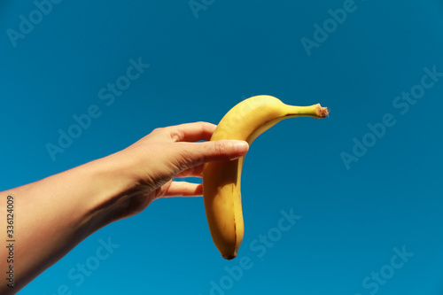 Imagen de una mano humana sujetando una banana amarilla sobre fondo azul del cielo