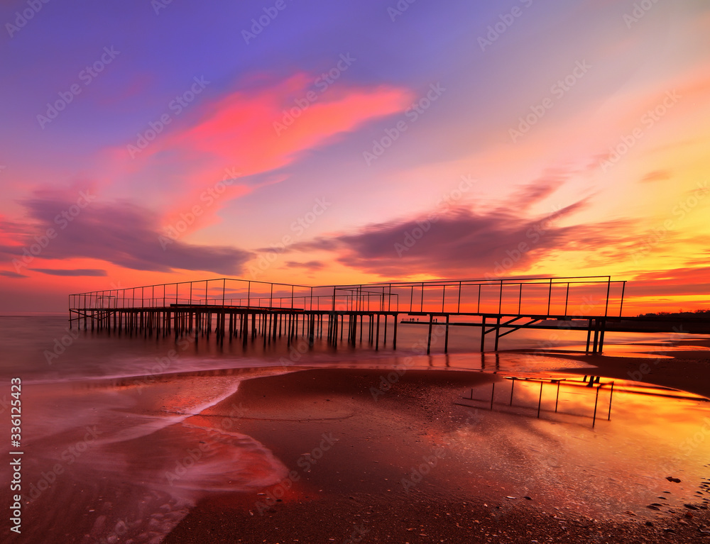 Fantastic sunset on a secluded beach. Old pier and reflection of red clouds in the water.
