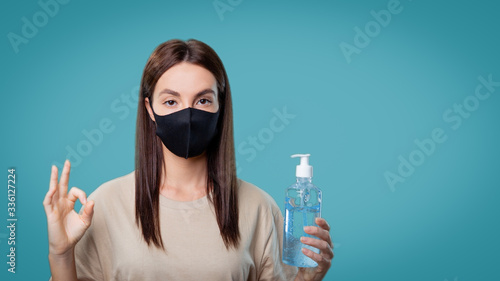Woman in medical mask holds a sanitizer and shows ok sign, isolated over blue