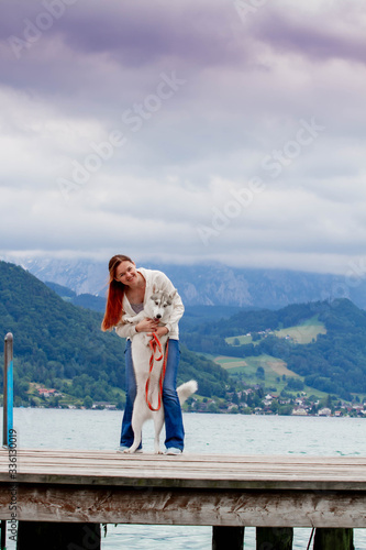 A young woman with brown hair and white sweater is sitting at the pier at the lake with calm water. A Siberian husky female dog is lying down near the girl. The Alps mountains in the background. © voltgroup