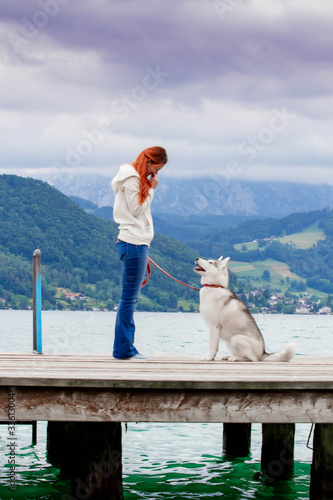 A young woman with brown hair and white sweater is sitting at the pier at the lake with calm water. A Siberian husky female dog is lying down near the girl. The Alps mountains in the background.