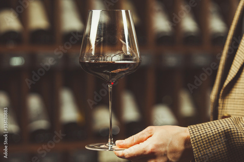 Glass of red wine in the sommelier hand on the cellar with bottles backgrounds.