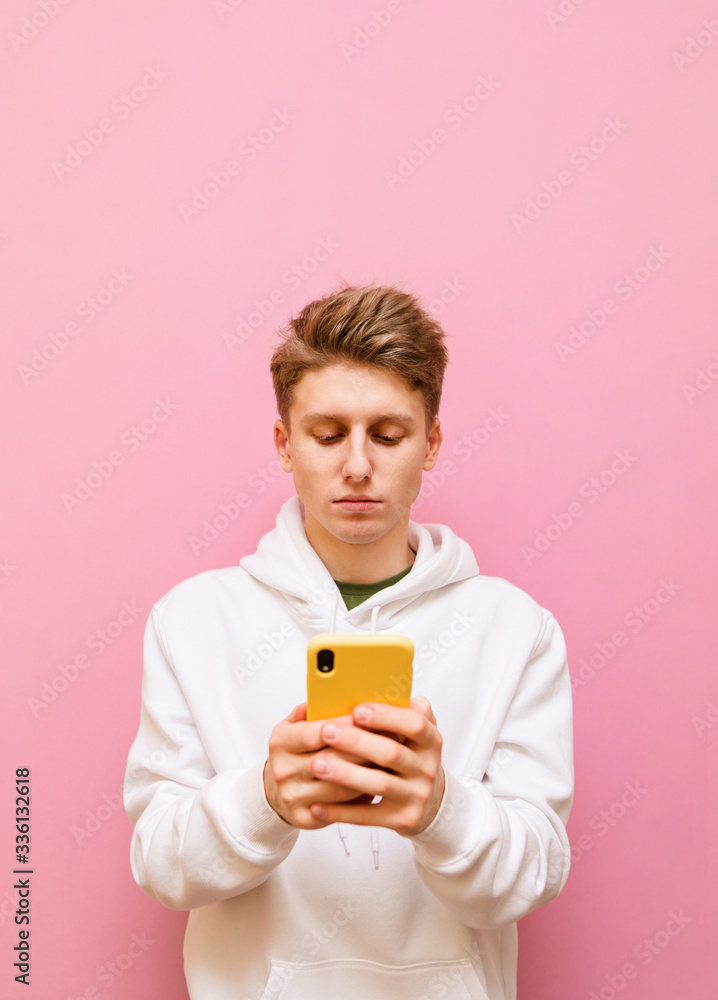 Portrait of a concentrated young man in a white hoodie stands on a pink background with a smartphone in his hands, looks intently into the screen. Guy student uses internet on smartphone, isolated. Portrait of a concentrated young man in a white hoodie stands on a pink background with a smartphone in his hands, looks intently into the screen. Guy student uses internet on smartphone, isolated.