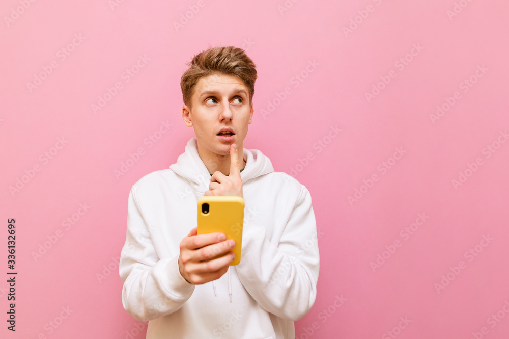 Portrait of thoughtful guy in white hoodie stands on pink background with smartphone in hands and looks away with pensive face. Teenager uses a smartphone, looks at copy space and thinks. Portrait of thoughtful guy in white hoodie stands on pink background with smartphone in hands and looks away with pensive face. Teenager uses a smartphone, looks at copy space and thinks.