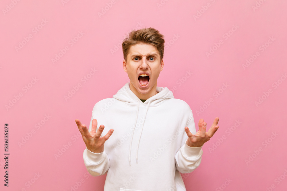 Angry young man in white casual clothes stands on a pink background and is nervous looking into the camera. Aggressive guy in white sweater isolated on pink background. Copy space Angry young man in white casual clothes stands on a pink background and is nervous looking into the camera. Aggressive guy in white sweater isolated on pink background. Copy space