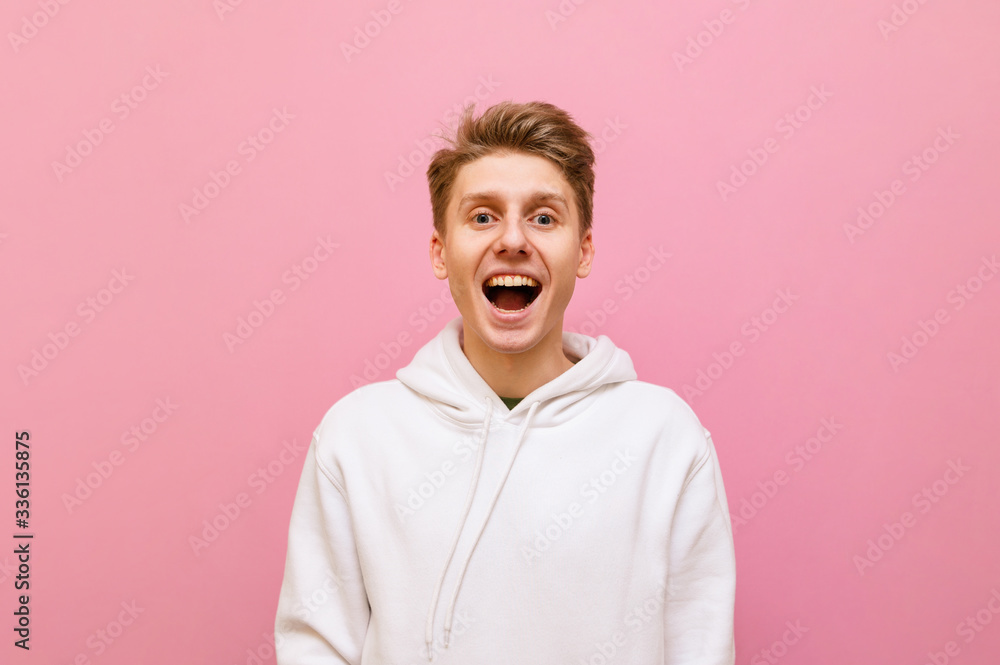 Portrait of joyful blond boy in white casual clothes stands on a pink background with his mouth open happily looking into the camera. Happy young man in white hoodie joyful posing on pink background. Portrait of joyful blond boy in white casual clothes stands on a pink background with his mouth open happily looking into the camera. Happy young man in white hoodie joyful posing on pink background.